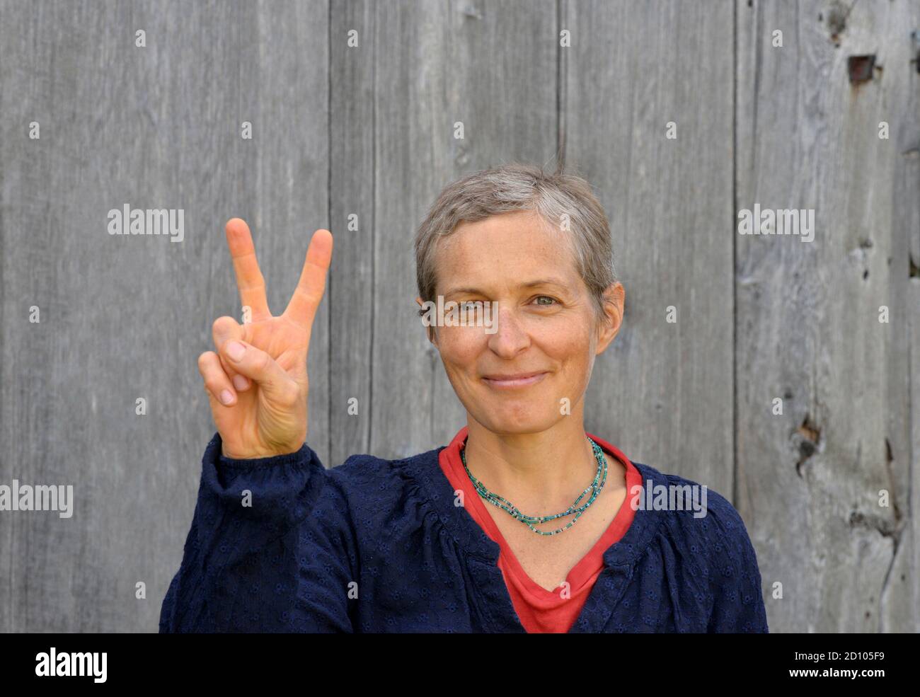 Erfolgreiche kaukasische Landfrau mittleren Alters mit kurzen Haaren zeigt das V-Zeichen (Sieg) mit der rechten Hand, vor altem Scheunenholzhintergrund. Stockfoto