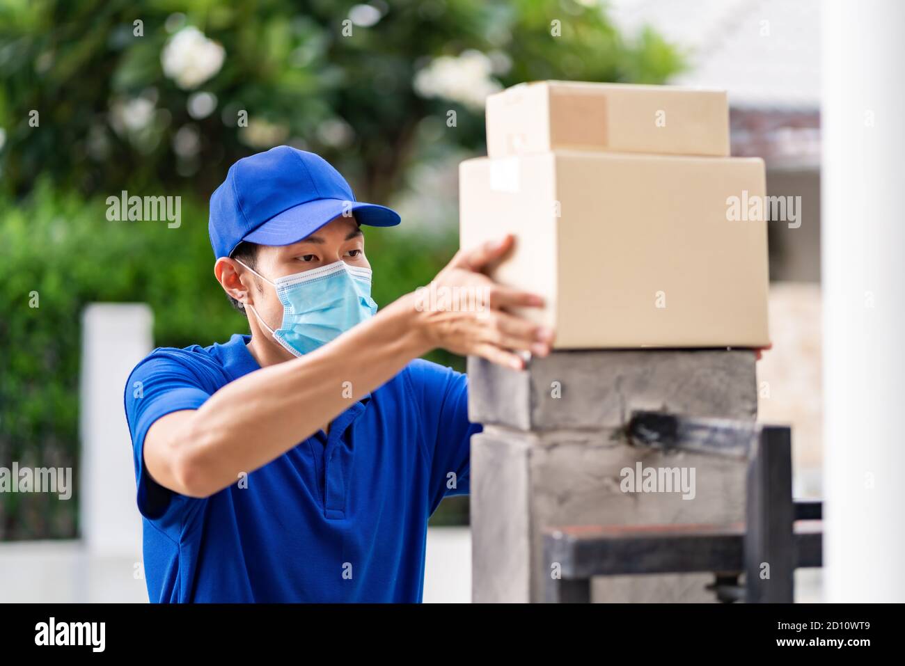 Asian liefern Mann mit Gesichtsmaske in blauen Shirt Handling-Pakete und setzen auf die Zaunpfosten des Kundenhauses als kontaktlose Shopping-Lieferung. Dies Stockfoto