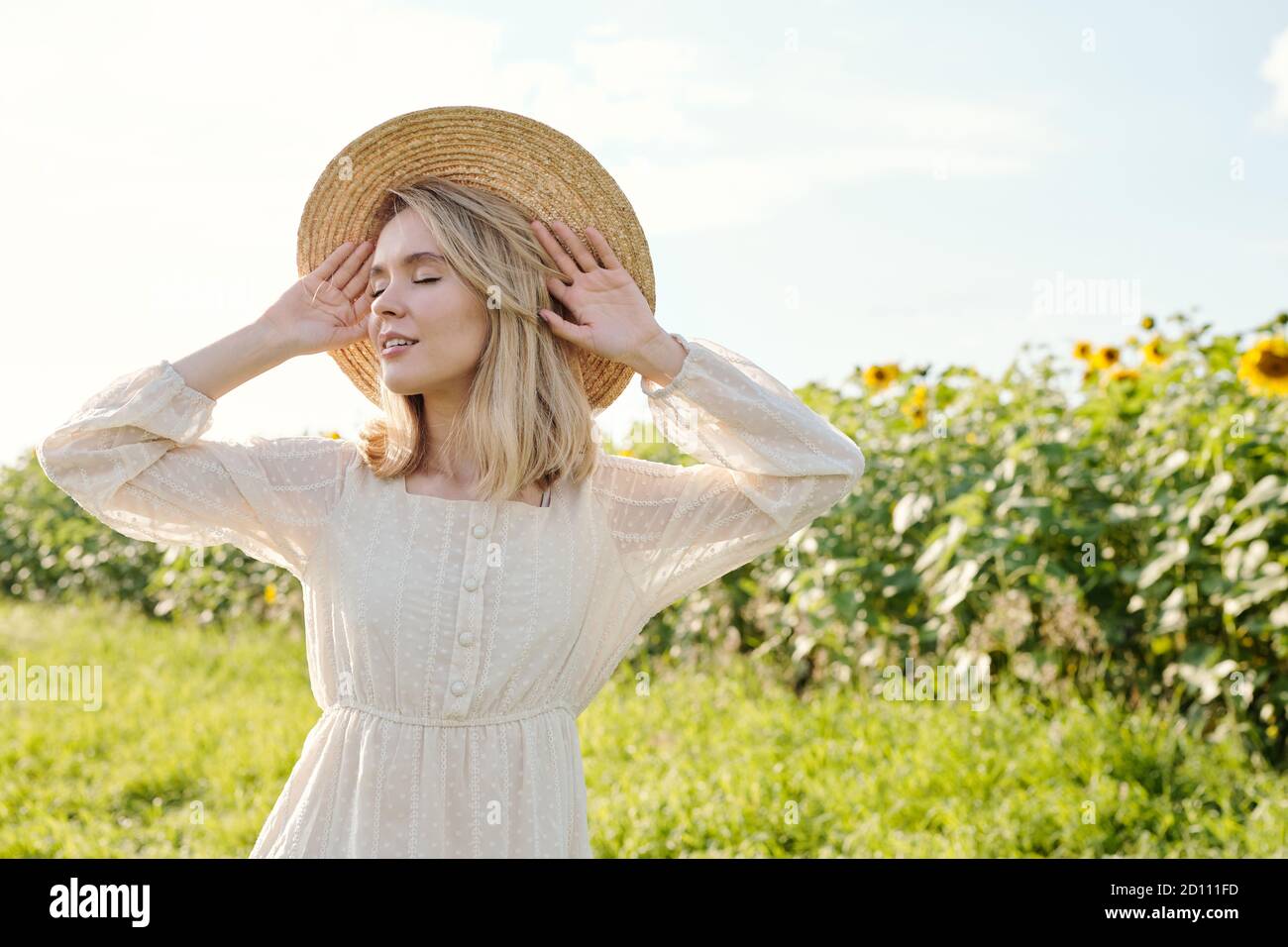 Schöne junge blonde Frau in Strohhut und weißem Kleid Augen geschlossen halten Stockfoto