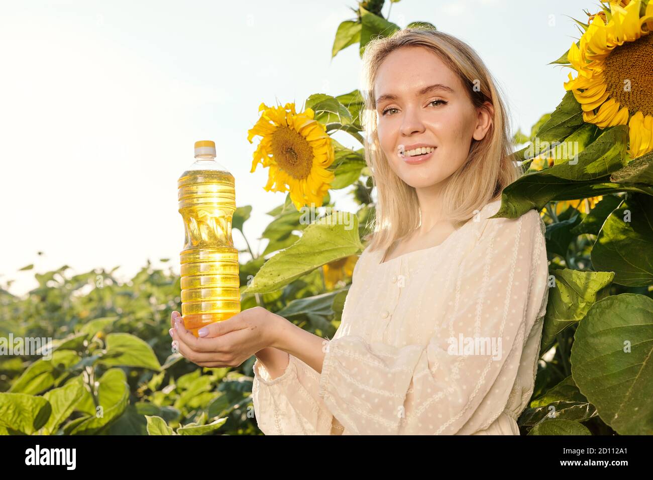 Lächelnd blonde junge Frau in weißem Kleid hält Flasche Sonnenblumenöl Stockfoto