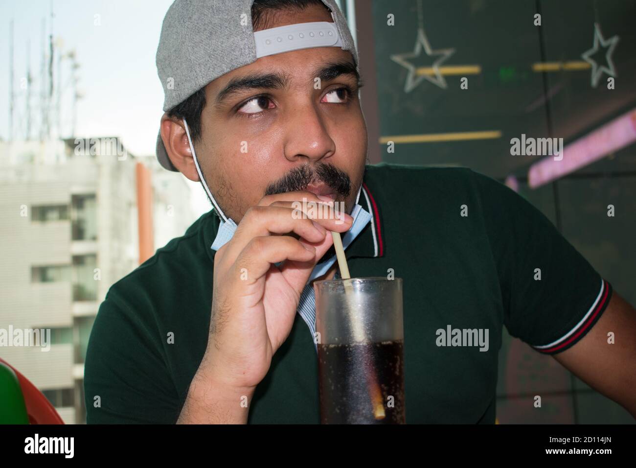 Schnurrbärte junger Mann mit Maske und Mütze in neue Normalität von CORONAVIRUS trinken mit Stroh und suchen mit Augen in einem Restaurant, Polo Green Shirt und essen Stockfoto