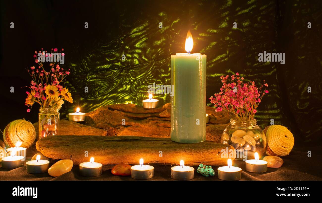 Komposition mit großer grüner Kerze umgeben von weißen Kerzen, Blumen und Steinen auf schwarzem Hintergrund mit grünem Licht Stockfoto