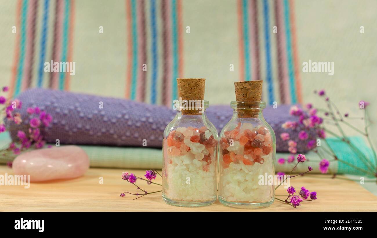 Zusammensetzung mit zwei Glasflaschen mit Badesalz auf Holztisch mit Handtüchern, rosa Stein, lila Blumen und Tuch mit Linien im Hintergrund Stockfoto