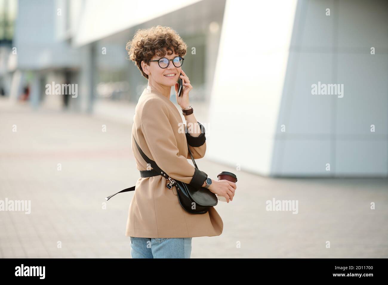 Zeitgenössische junge Geschäftsfrau mit Getränk und Handtasche per Smartphone Stockfoto
