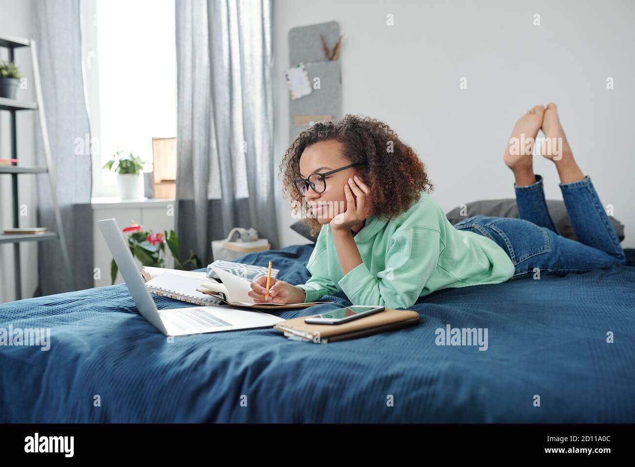 Junge clevere Mixed-Race-Schüler beobachten Online-Unterricht beim Notizen machen Stockfoto