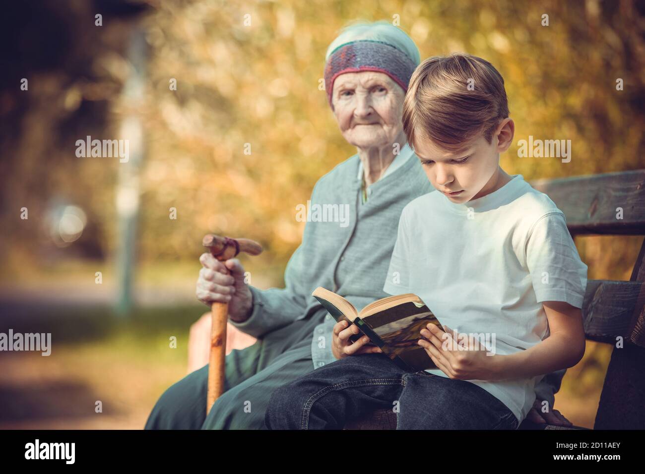 Kleiner Junge, der Buch auf der Bank im Park liest. Seine Urgroßmutter sitzt neben ihm. Stockfoto