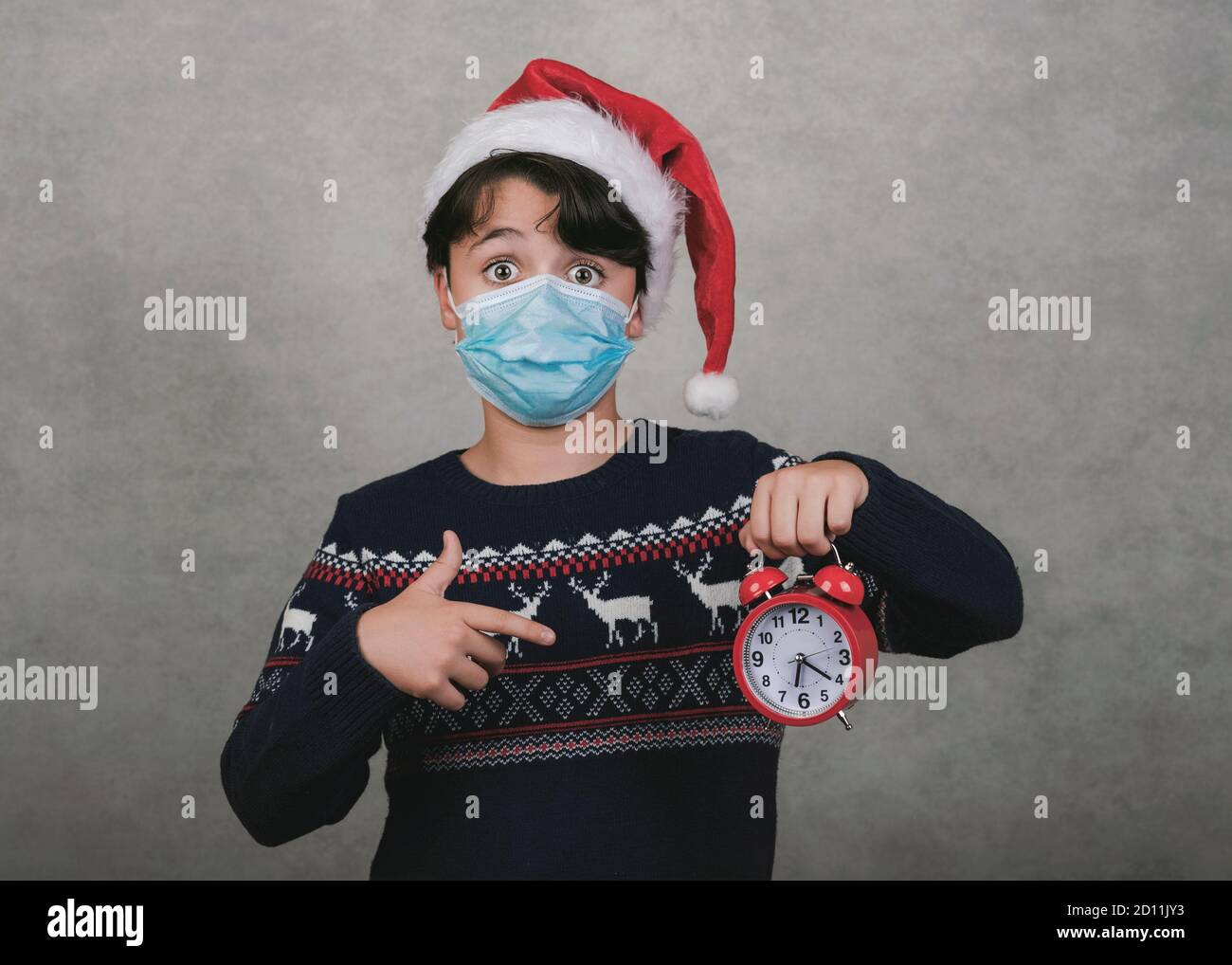 Frohe Weihnachten, Lustiges Kind mit medizinischer Maske und Wecker trägt Weihnachtsmann Hut auf grauem Hintergrund Stockfoto