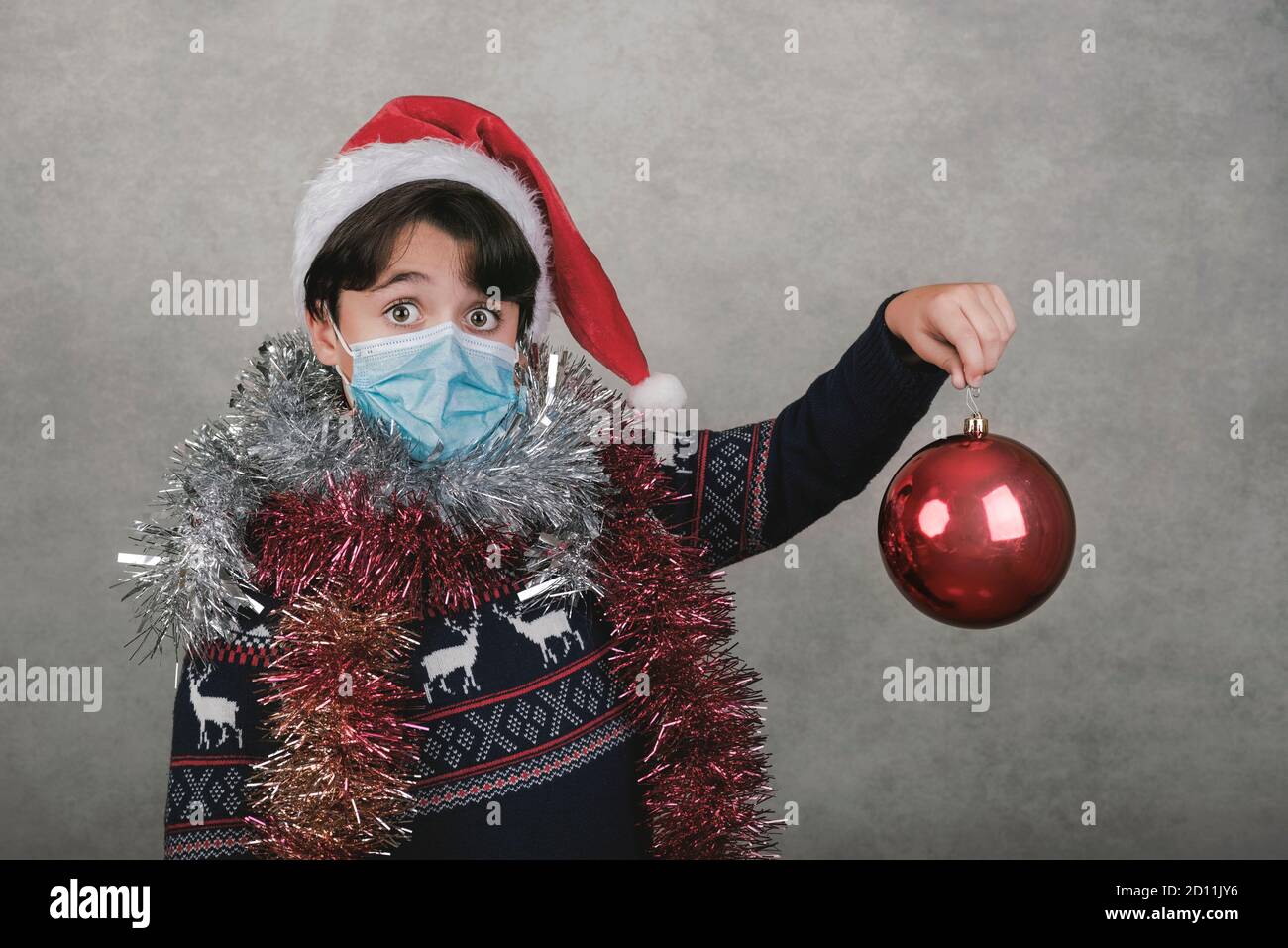 Frohe Weihnachten, Junge mit medizinischer Maske halten eine weihnachtskugel über grauem Hintergrund Stockfoto