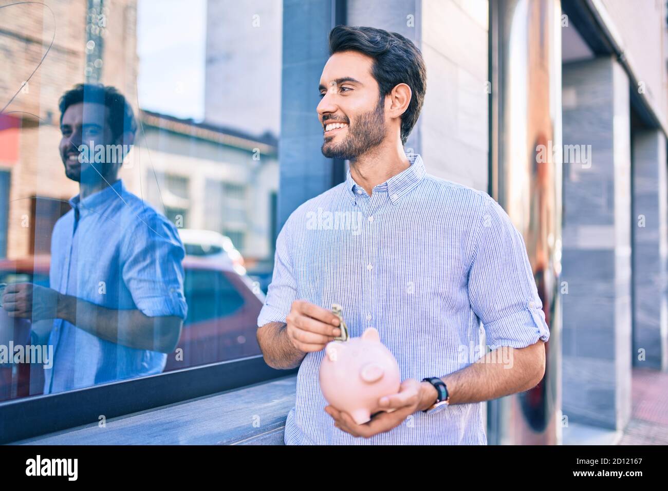 Junger hispanischer Mann lächelt glücklich Speichern Banknote im Sparschwein in der Stadt. Stockfoto