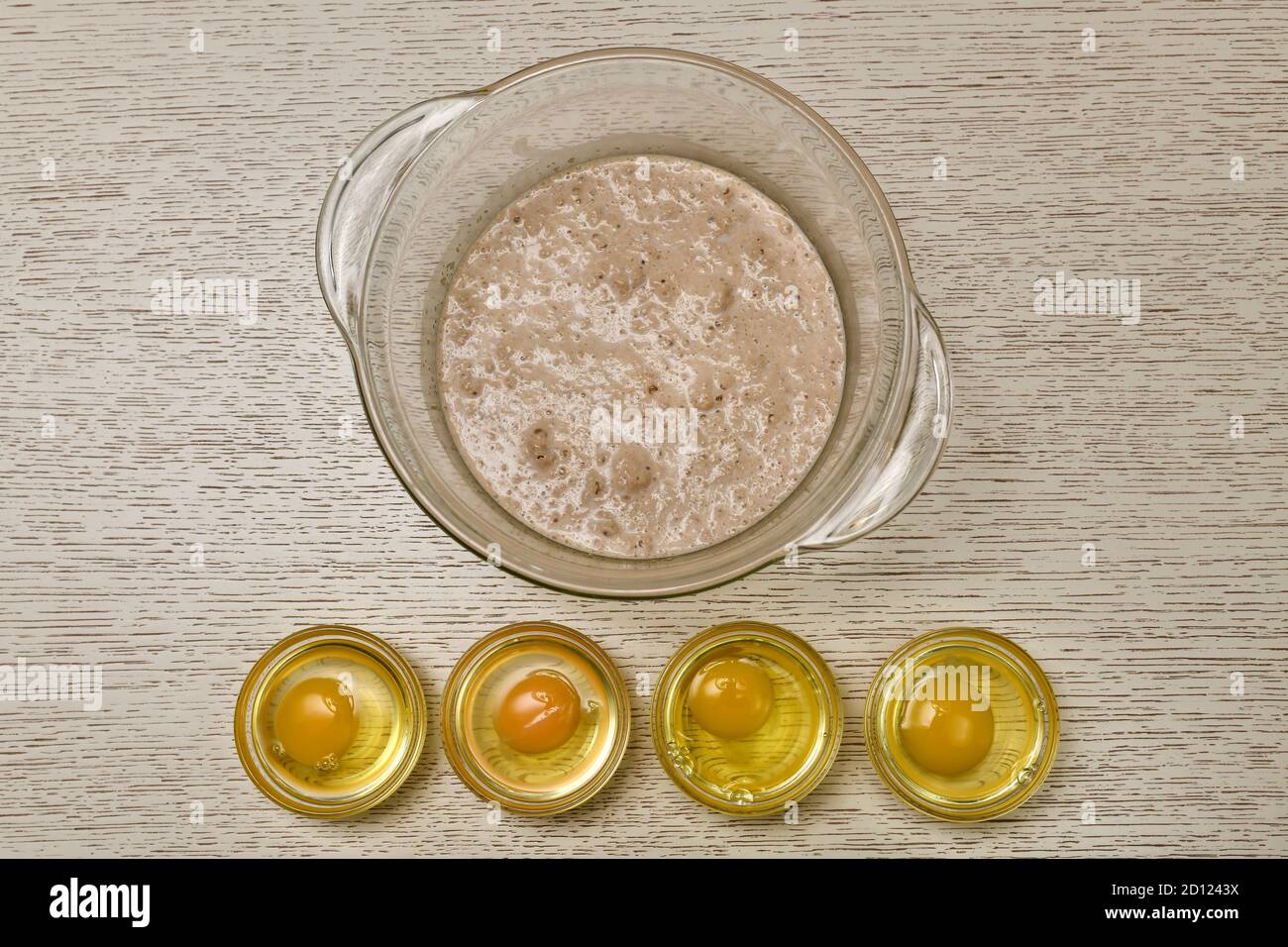 Die Stufe der Vorbereitung von Eiern für das Kneten von Hefeteig. Tiefes Glasgefäß mit Hefe in Milch. Vier rohe Eier in einer Schüssel. Stockfoto