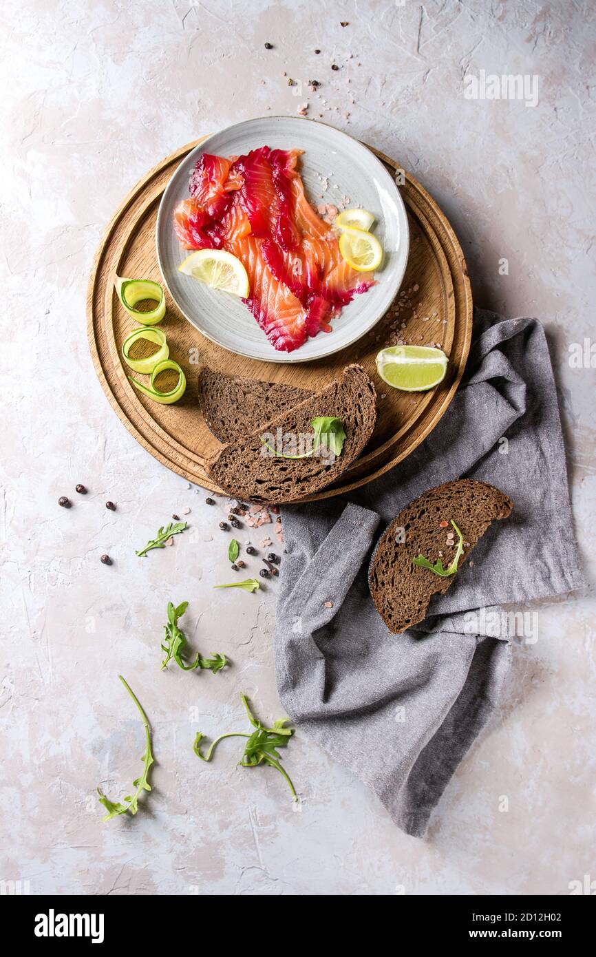 In Scheiben geschnittene rote Beete marinierter Lachs für Sandwiches mit in Scheiben geschnittenen Roggenbrot, rosa Salz, Pfeffer, grün, Gurke, Zitrone auf Holz- rundes Tablett serviert auf texti Stockfoto