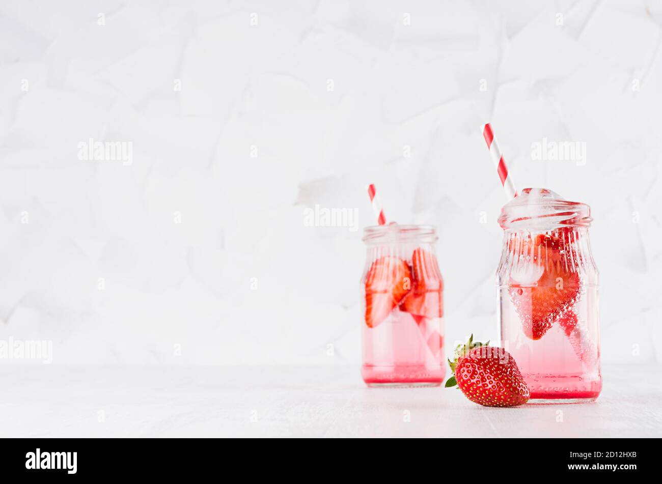 Natürliche Bio-Cocktails mit Erdbeere, kohlensäurehaltiges Wasser und Eiswürfeln mit gestreiftem Stroh auf hellweißem Holzbrett und Wand, Kopierraum. Stockfoto
