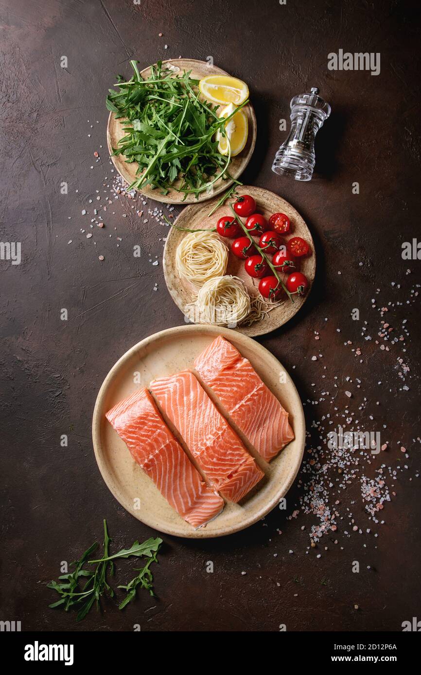 Geschnittenem rohen ungekochten Lachs filet auf Keramikteller mit Zutaten für Abendessen, Rucola, Zitrone, Salz, Pfeffer, Nudeln, Cherry Tomaten über Dunkelbraun Stockfoto