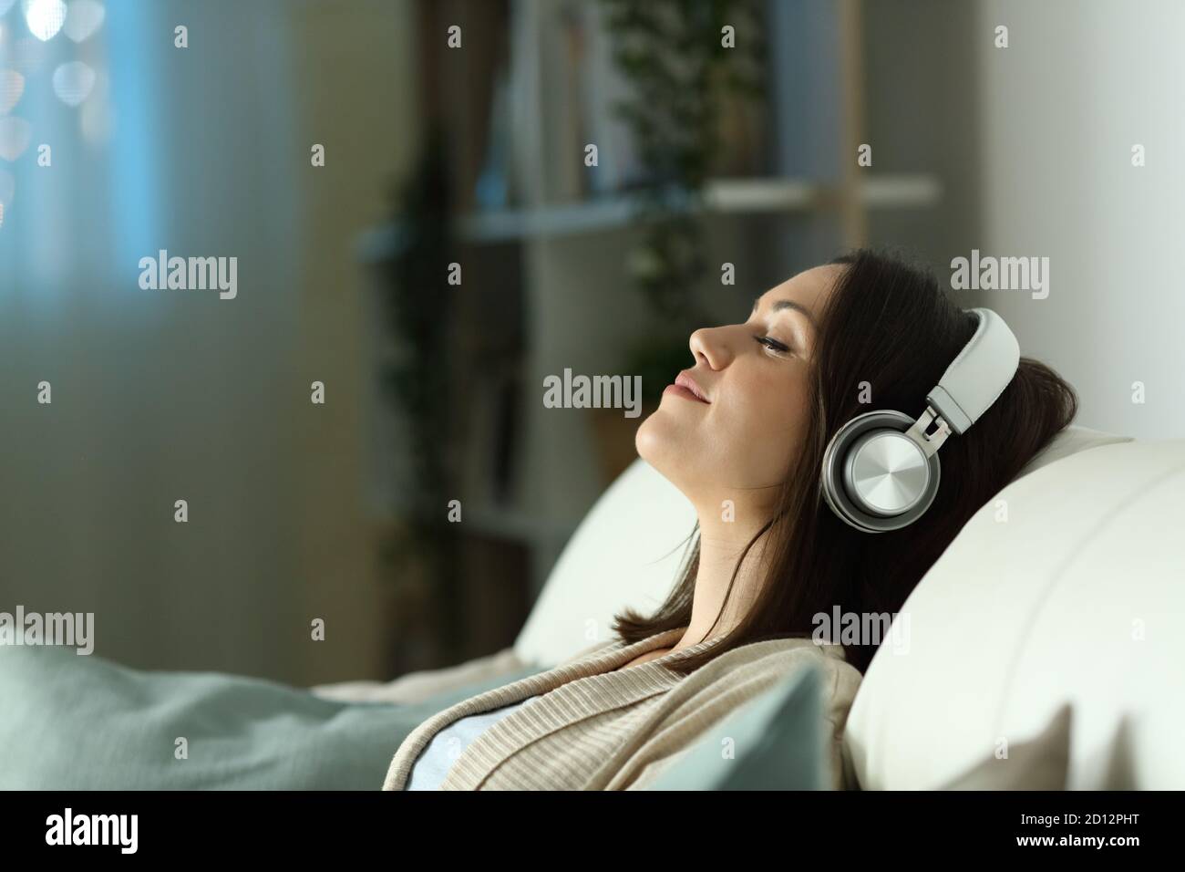 Profil einer entspannten Frau, die mit Kopfhörern Musik hört In der Nacht auf einer Couch im Wohnzimmer sitzen Zimmer zu Hause Stockfoto