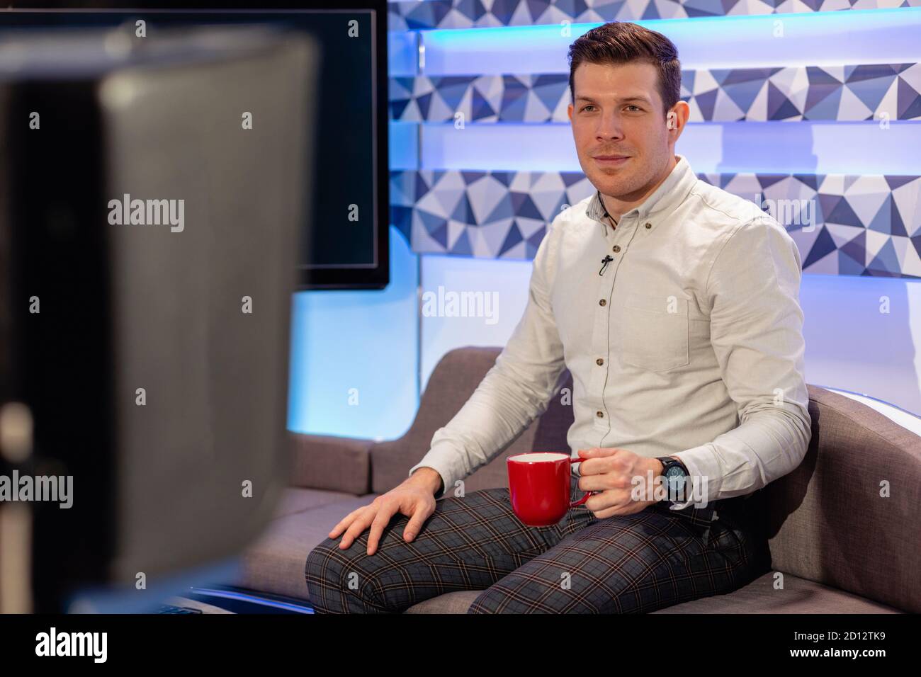 Ein Fernsehmoderator sitzt auf einem Sofa in einem Filmstudio und hält eine Tasse Tee. Er macht eine Pause vom Filmen einer TV-Show. Stockfoto