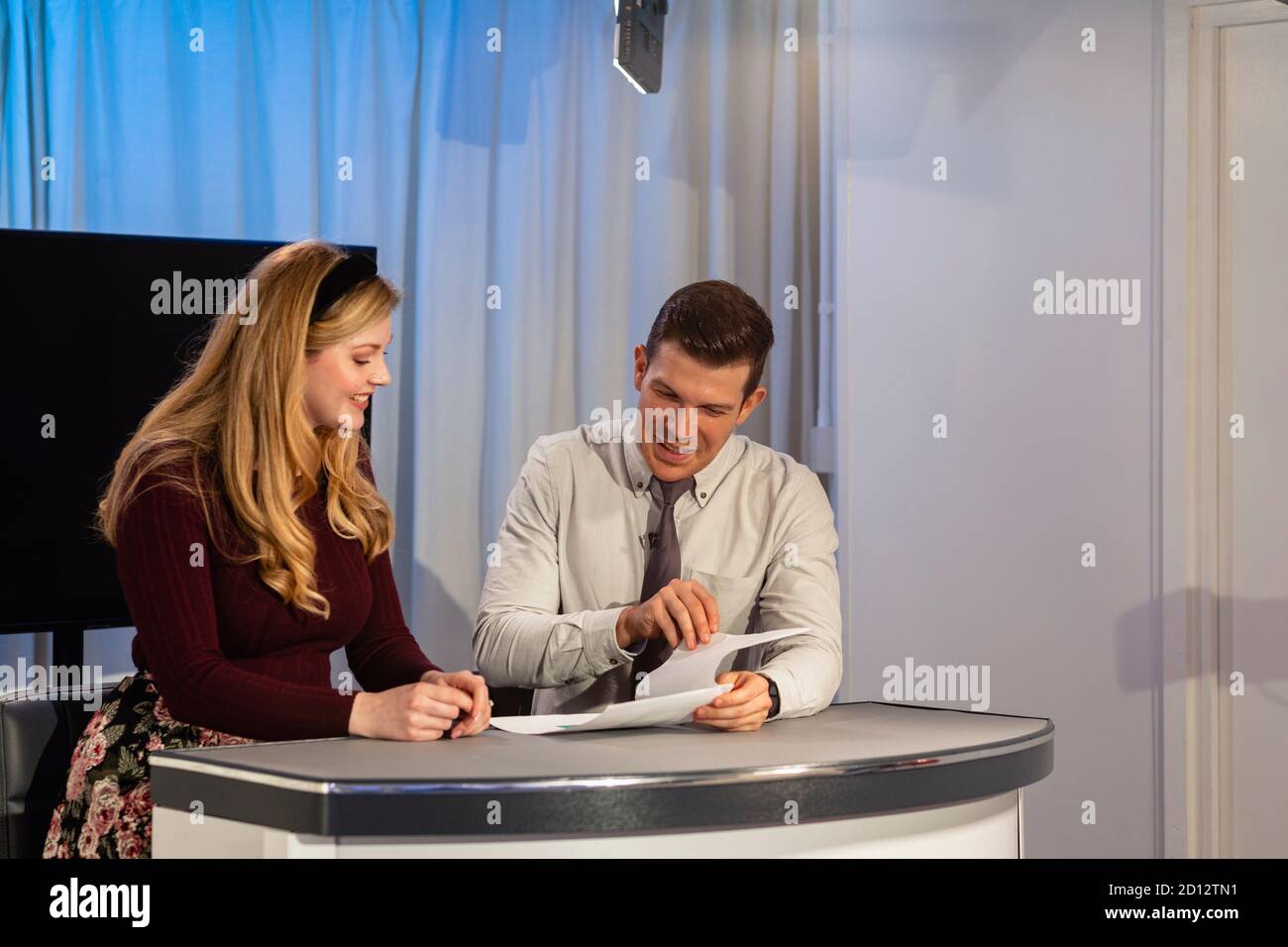 Zwei Fernsehmoderatoren sitzen an einem Schreibtisch in einem Filmstudio. Sie lesen die Show-Notizen zusammen und bereiten sich darauf vor, wieder zu Filmen. Stockfoto