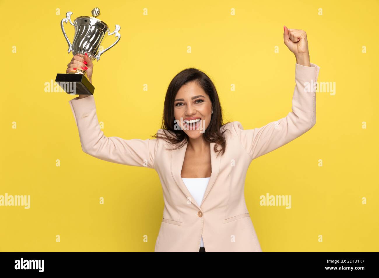 Fröhliche junge Frau hält Fäuste in der Luft und feiert den Sieg, hält silberne Trophäe und lacht auf gelbem Hintergrund Stockfoto