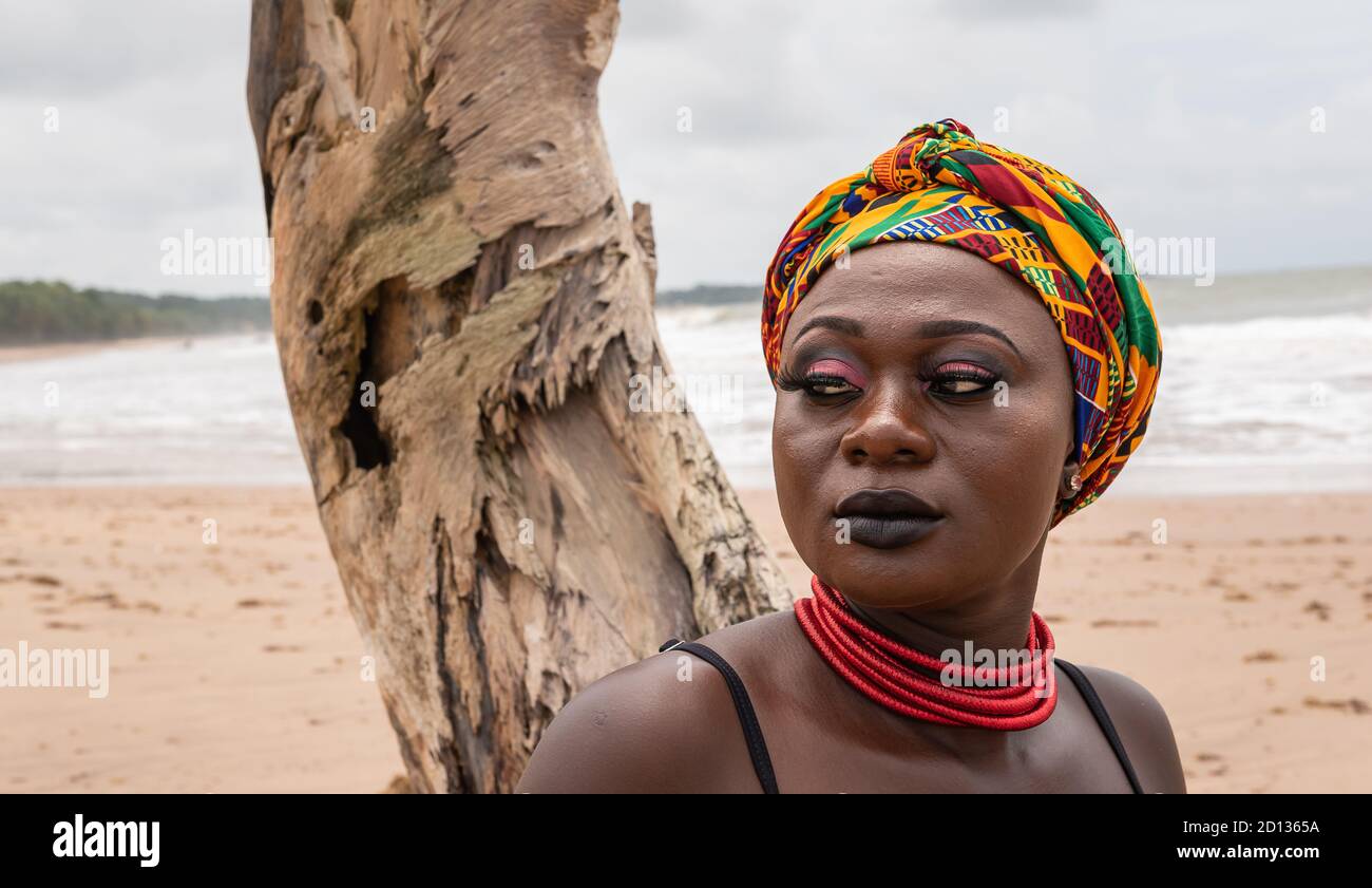 Ghanaische afrikanische Frau mit buntem Kopfschmuck sitzt an einem gebrochenen Palmenstumpf am Strand in Axim Ghana West Afrika Stockfoto