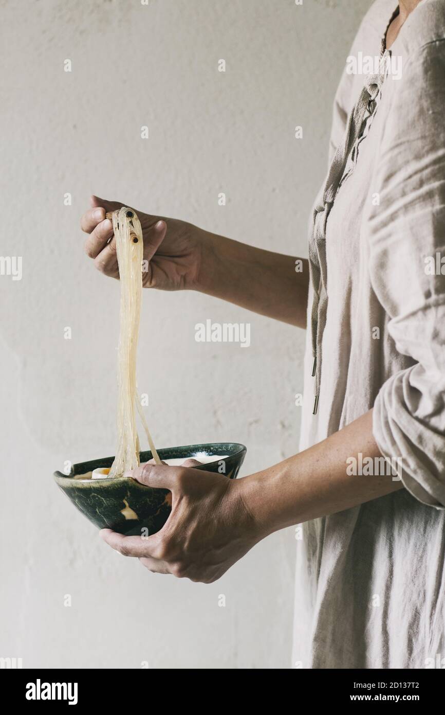 Frau in weißem Kleid Holding in den Händen Schüssel der Asiatischen Reis und Nudeln Nudelsuppe mit Stäbchen. Graue Wand als Hintergrund. Abendessen im asiatischen Stil. Stockfoto