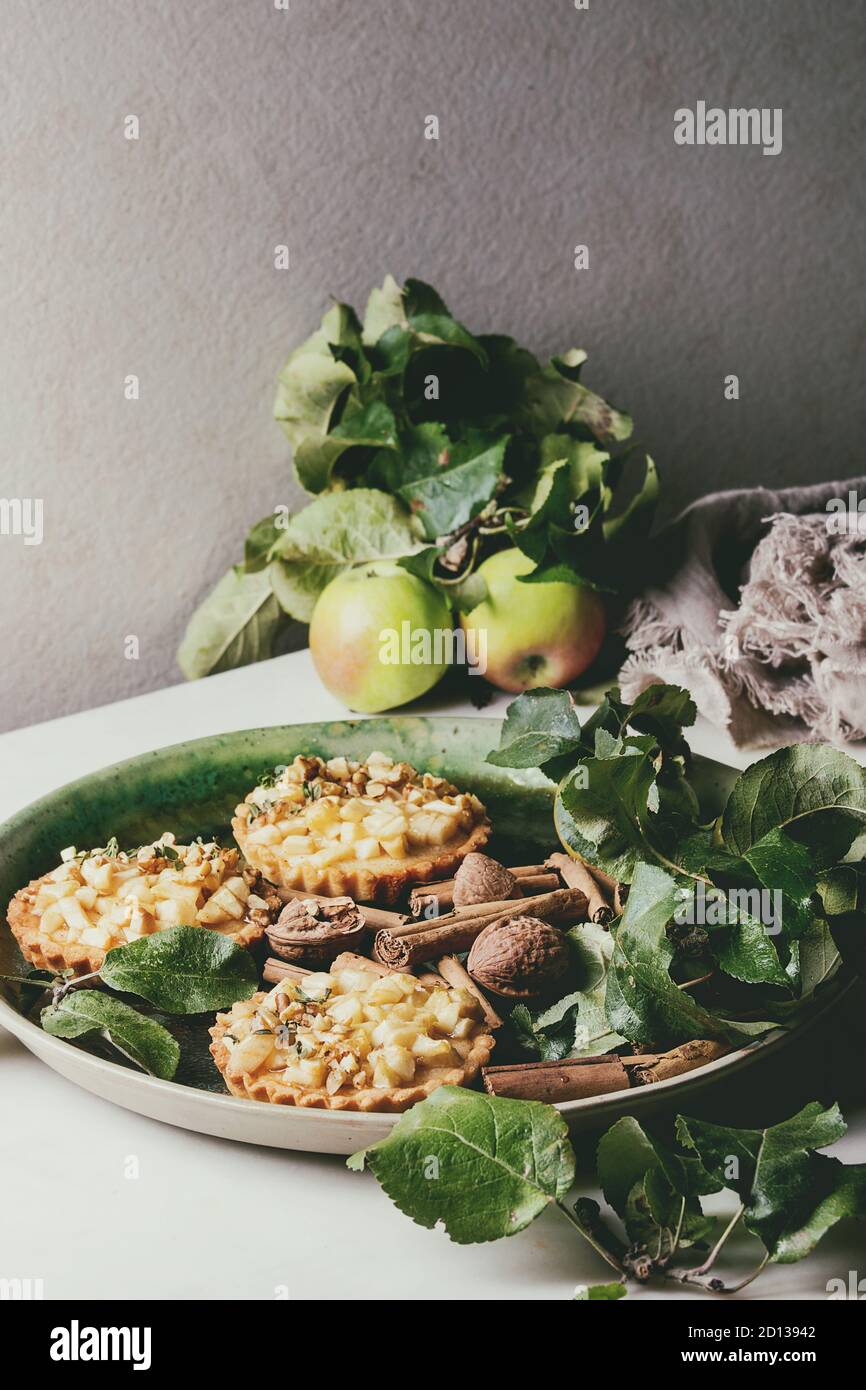 Hausgemachten süßen Apple shortbread Törtchen in grüner Keramikteller mit Zimtstangen, Walnüsse, Äpfel Zweige oben auf weißem Marmortisch. Herbst bak Stockfoto