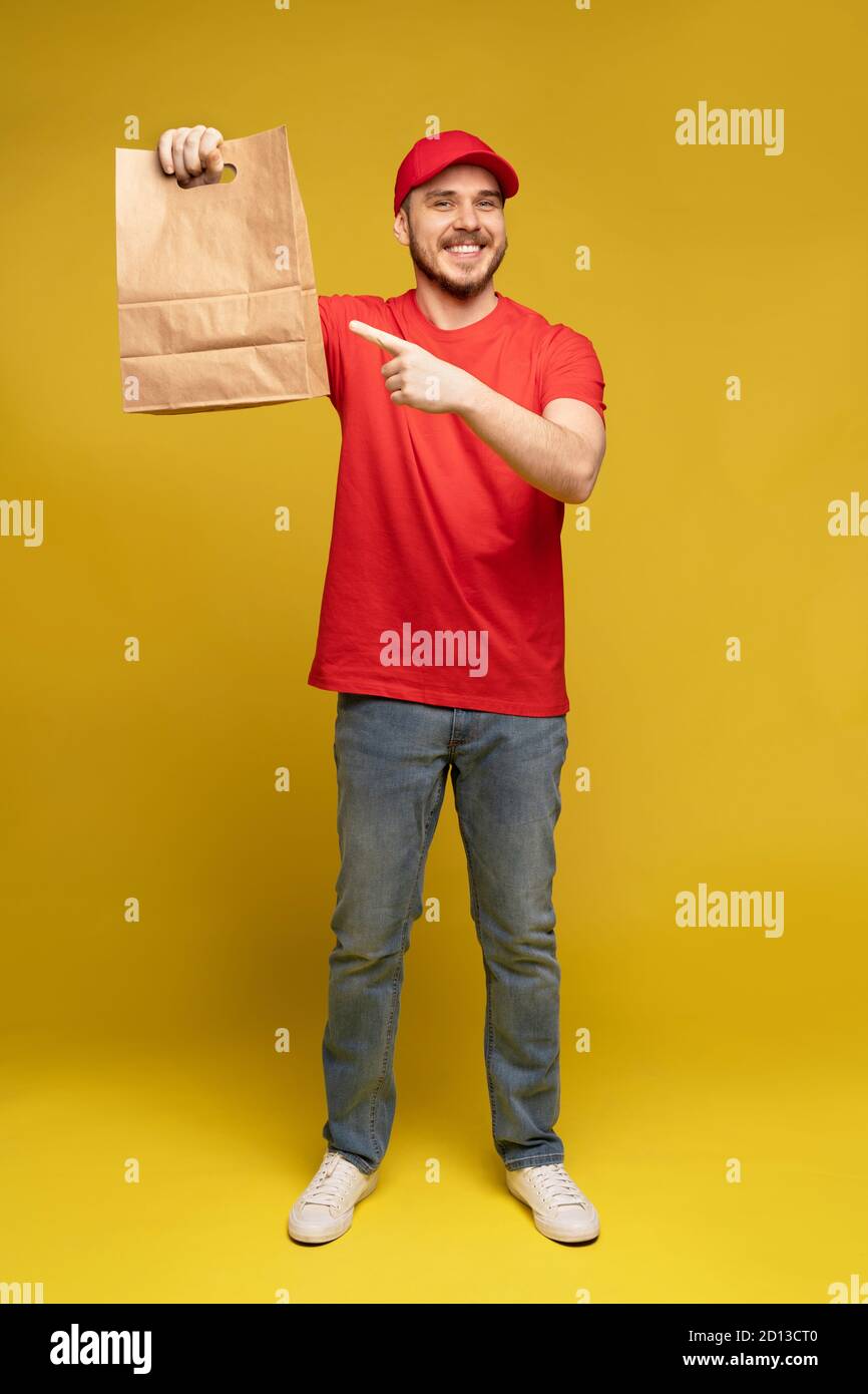 Mann in roter Mütze, T-Shirt mit Fast-Food-Bestellung isoliert auf gelbem Hintergrund. Männlicher Mitarbeiter Kurier halten leere Papierpaket mit Essen Stockfoto