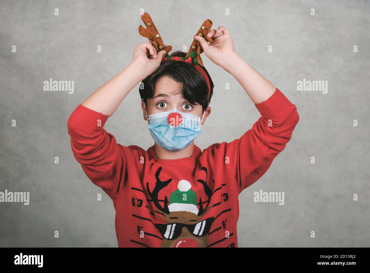 Frohe Weihnachten, lustiges Kind mit medizinischer Maske in einem Rudolph Rentier weihnachtskostüm auf grauem Hintergrund Stockfoto