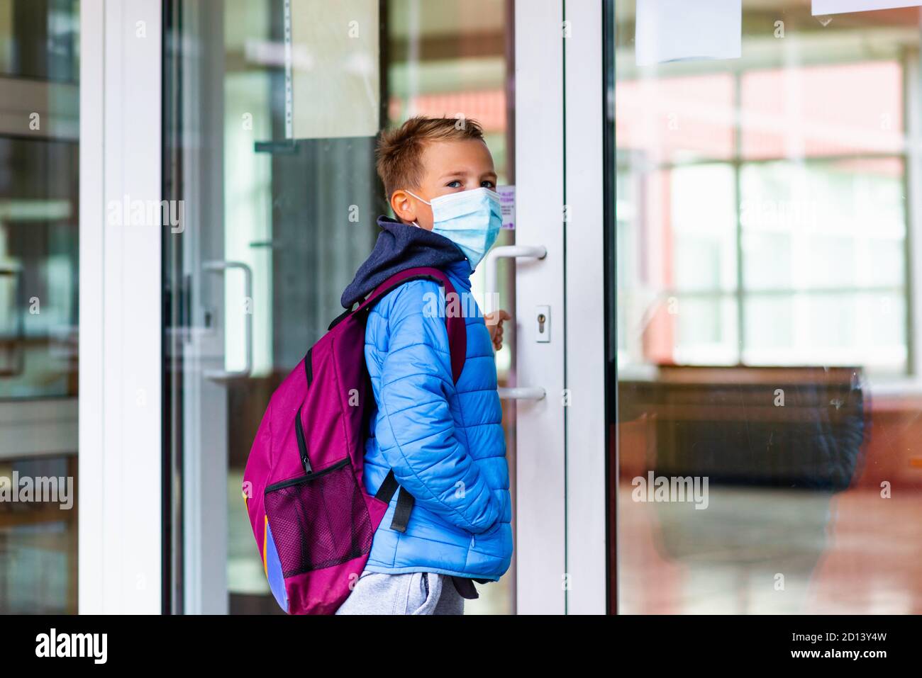 Junge Schuljunge trägt Schutzmaske versucht, die Schultür zu öffnen. Hinter dem Rucksack Schoolboy zur Seite schauen Stockfoto