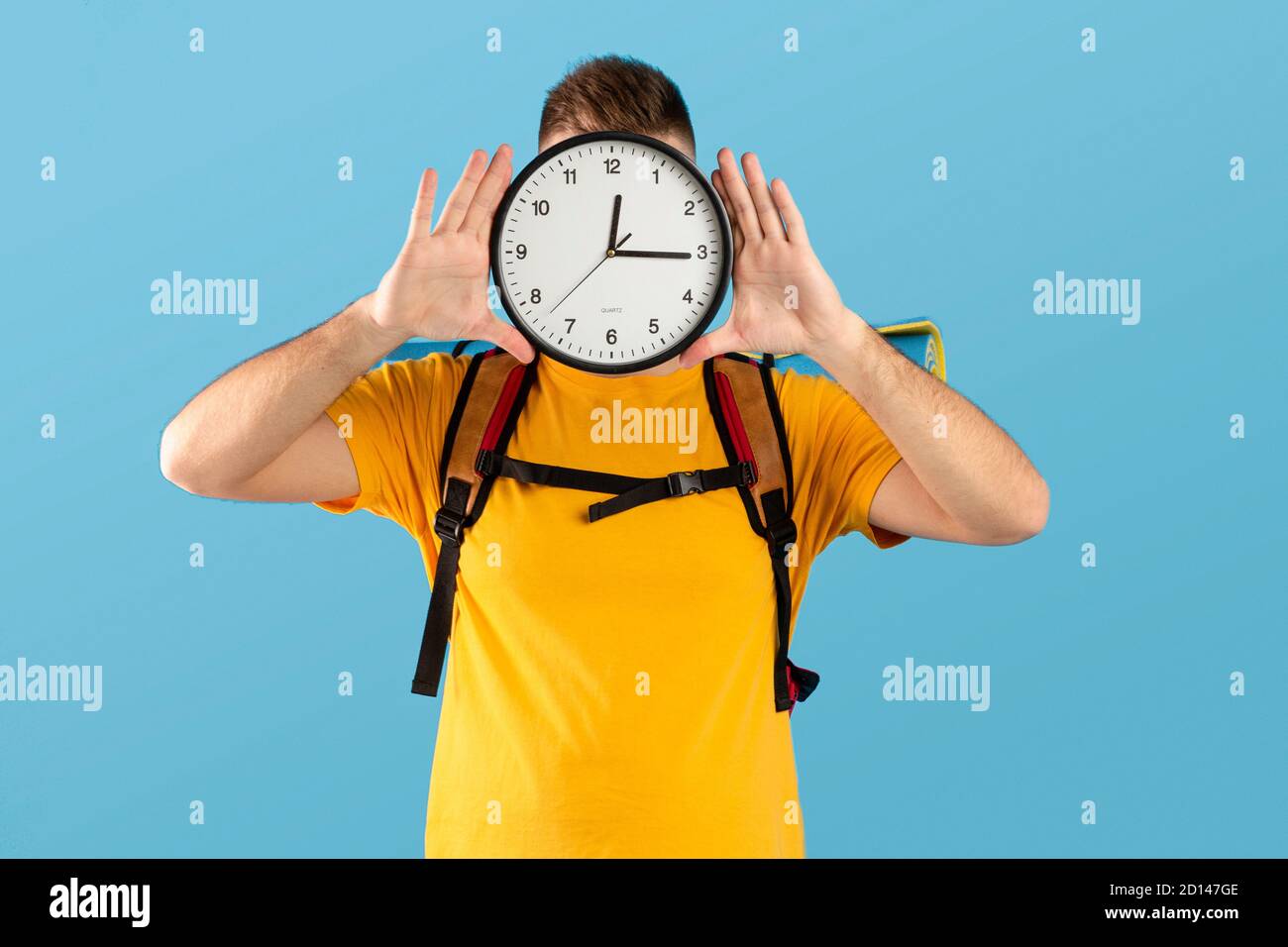 Time-to-Travel-Konzept. Junger Mann mit Campingausrüstung, die Uhr vor seinem Gesicht über blauem Studiohintergrund hält Stockfoto