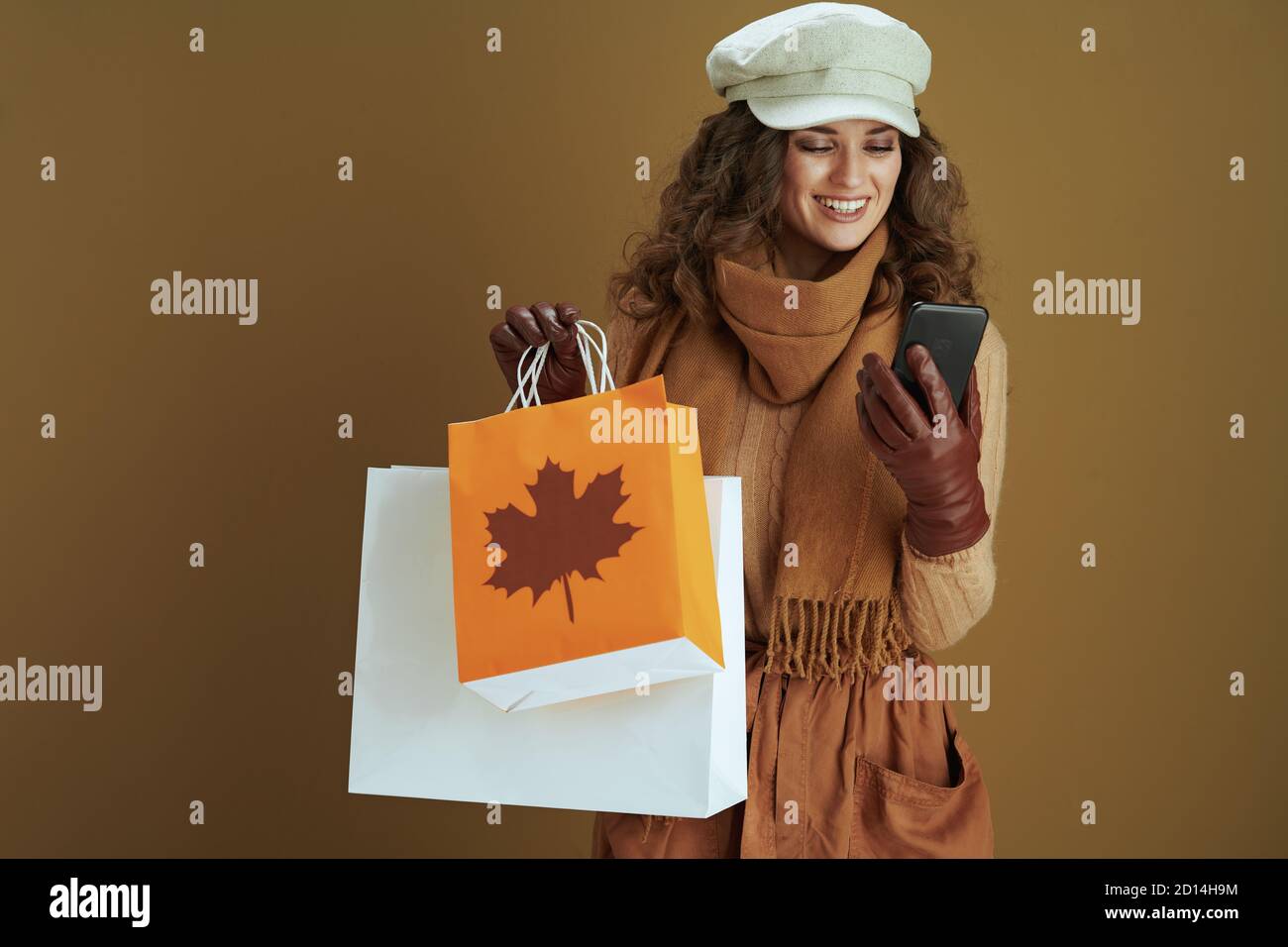Hallo Herbst. Lächelnd moderne 40 Jahre alte Hausfrau in Pullover mit Smartphone, Lederhandschuhe und Papier Einkaufstasche isoliert auf braunem Hintergrund. Stockfoto