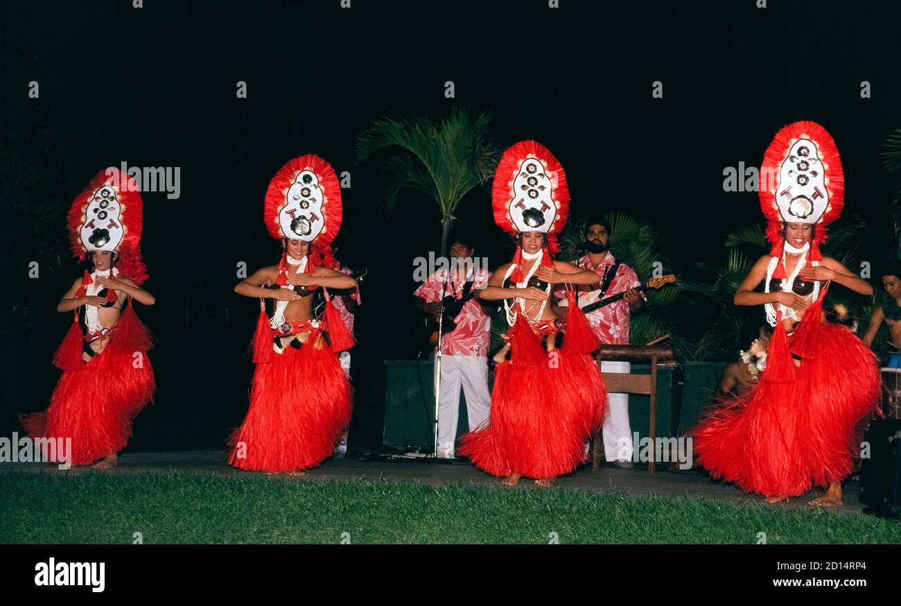 Vier lächelnde weibliche Entertainer treten in leuchtend roten Kostümen während eines Hawaii Luau auf, das einheimische Speisen und Tänze in einem Ferienresort auf der Insel Maui in Hawaii, USA, bietet. Ein Luau ist der beste Ort für Besucher, um traditionelle Hula-Tänze zu sehen, wo alte hawaiianische Geschichten mit den Händen und Armen der Tänzer erzählt werden. Tänze anderer polynesischer Kulturen sind oft Teil der Show, vor allem von Tahitian-otea-Tänzern, wie sie hier zu sehen sind. Sie sind leicht durch hohe aufwendige Kopfbedeckungen, Kokosnussschalen und große Grasröcke zu identifizieren, die mit den schnell schüttelnden Hüften der Mädchen vibrieren. Stockfoto