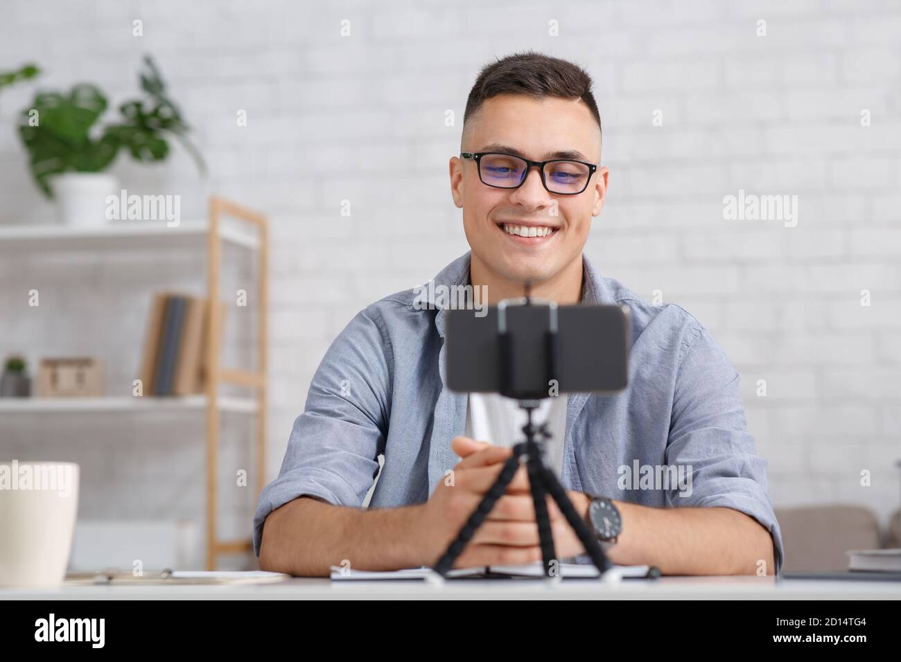 Moderne Arbeit von zu Hause aus während covid-19 Ausbruch. Lächelnder Mann mit Brille schaut auf das Smartphone auf dem Stativ und macht Aufnahme oder Videoanruf Stockfoto