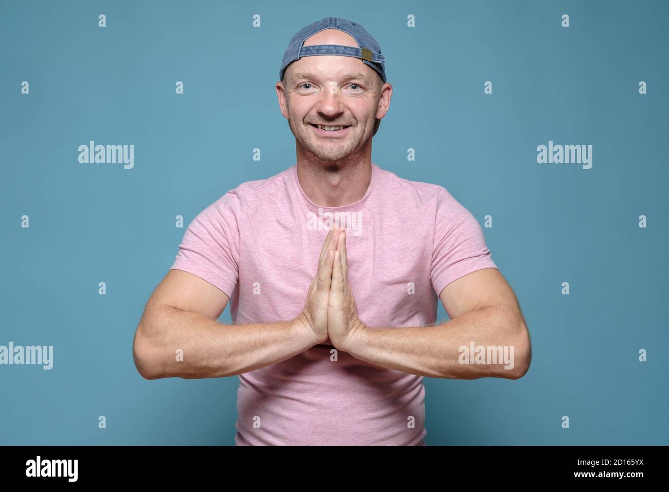 Wohlwollender, lächelnder Mann faltete die Hände in einer namastigen Geste. Blauer Hintergrund. Stockfoto