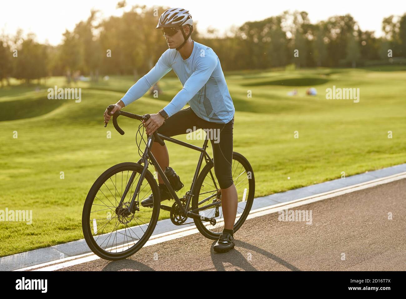 Sommerradtraining. Junger Athletic Mann in Sportbekleidung Fahrrad entlang einer Straße im Stadtpark bei Sonnenuntergang, voller Länge. Gesunder aktiver Lebensstil und Sportkonzept Stockfoto