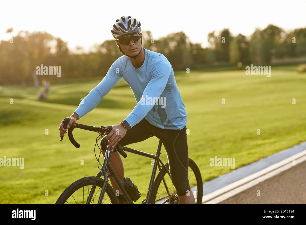 Starker Sportler in Sportbekleidung und Schutzhelm, der mit seinem Fahrrad auf der Straße im Park steht und wegschaut, nach dem Radfahren im Freien ausruhend. Gesunder aktiver Lebensstil und Sportkonzept Stockfoto