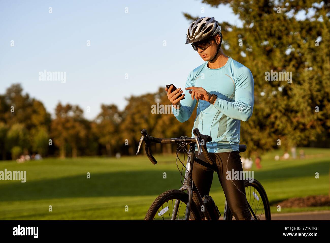 Ergebnisse werden überprüft. Junger Athlet, Profi-Radfahrer in Sportbekleidung und Schutzhelm stehen mit seinem Fahrrad im Park und mit seinem Smartphone. Aktiver Lebensstil und Sport Stockfoto