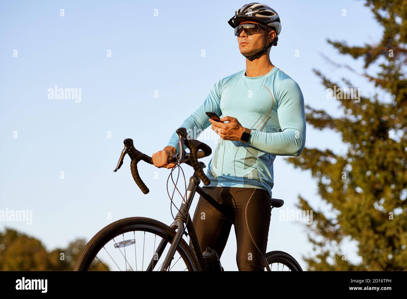 Profi-Radler in Sportbekleidung hält Smartphone und schaut weg, Ruhe nach der Fahrt, während er mit seinem Fahrrad im Park an einem sonnigen Tag stehen. Aktiver Lebensstil und Sport Stockfoto
