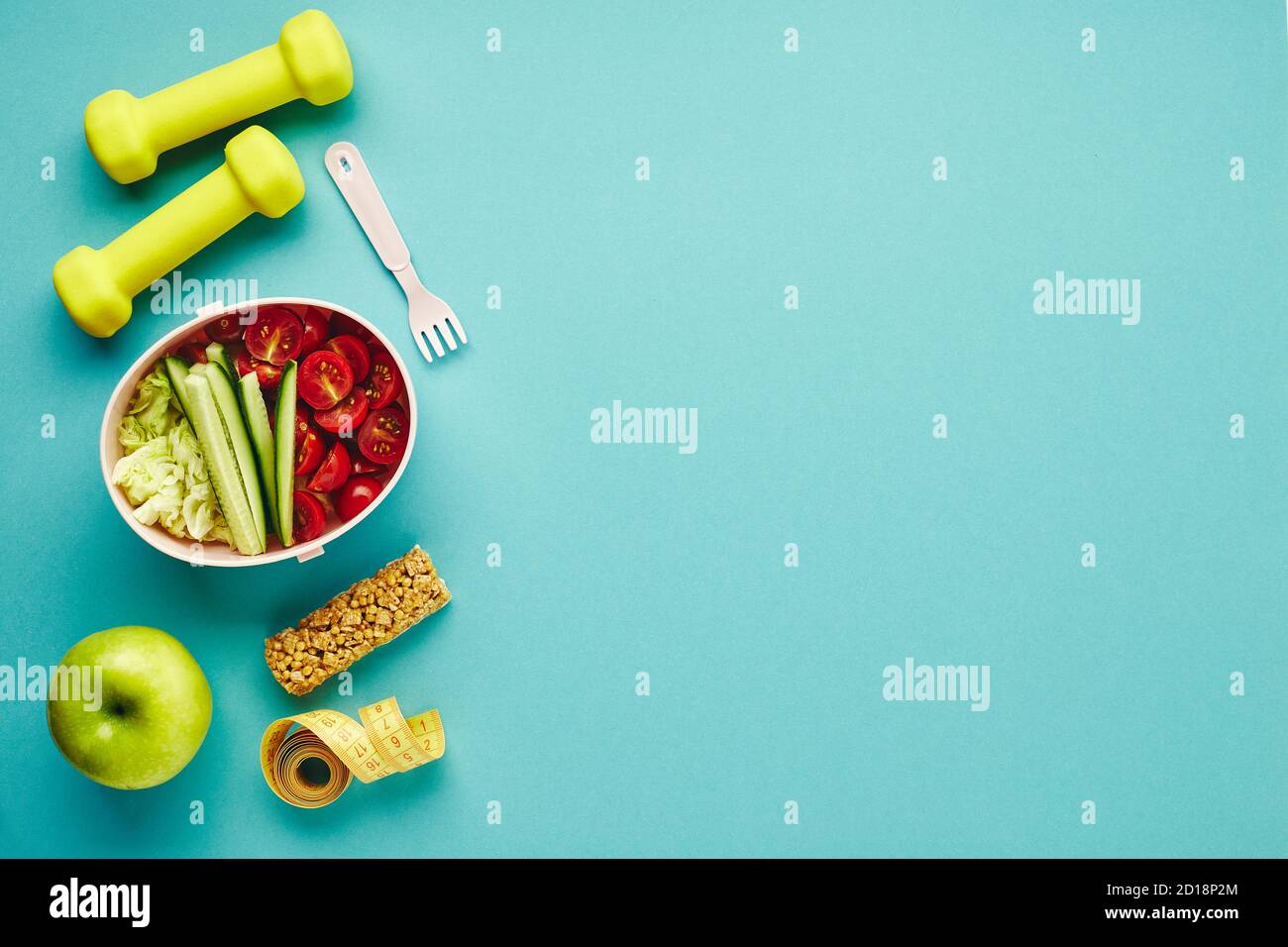 Kreative flache Lay von Sport- und Fitnessgeräten. Kurzhanteln, Maßband und Lunchbox mit gesundem Gemüsesalat auf hellblauem Hintergrund. Stockfoto