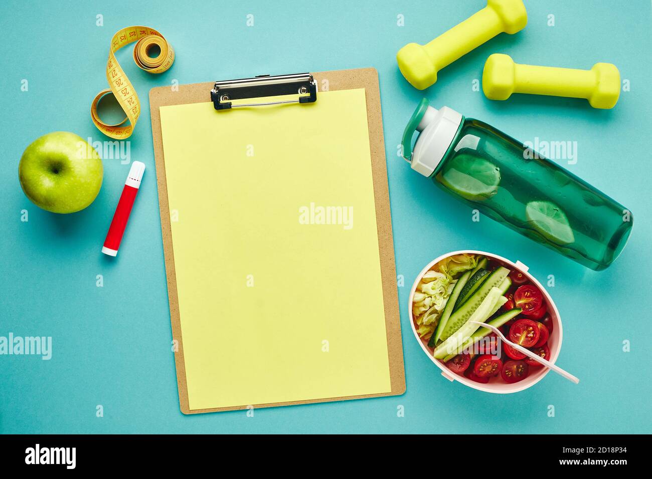 Falttablett für Papier, Wasserflasche, Kurzhanteln, Maßband und Lunchbox mit gesundem Gemüsesalat auf hellblauem Hintergrund. Das Konzept von Stockfoto