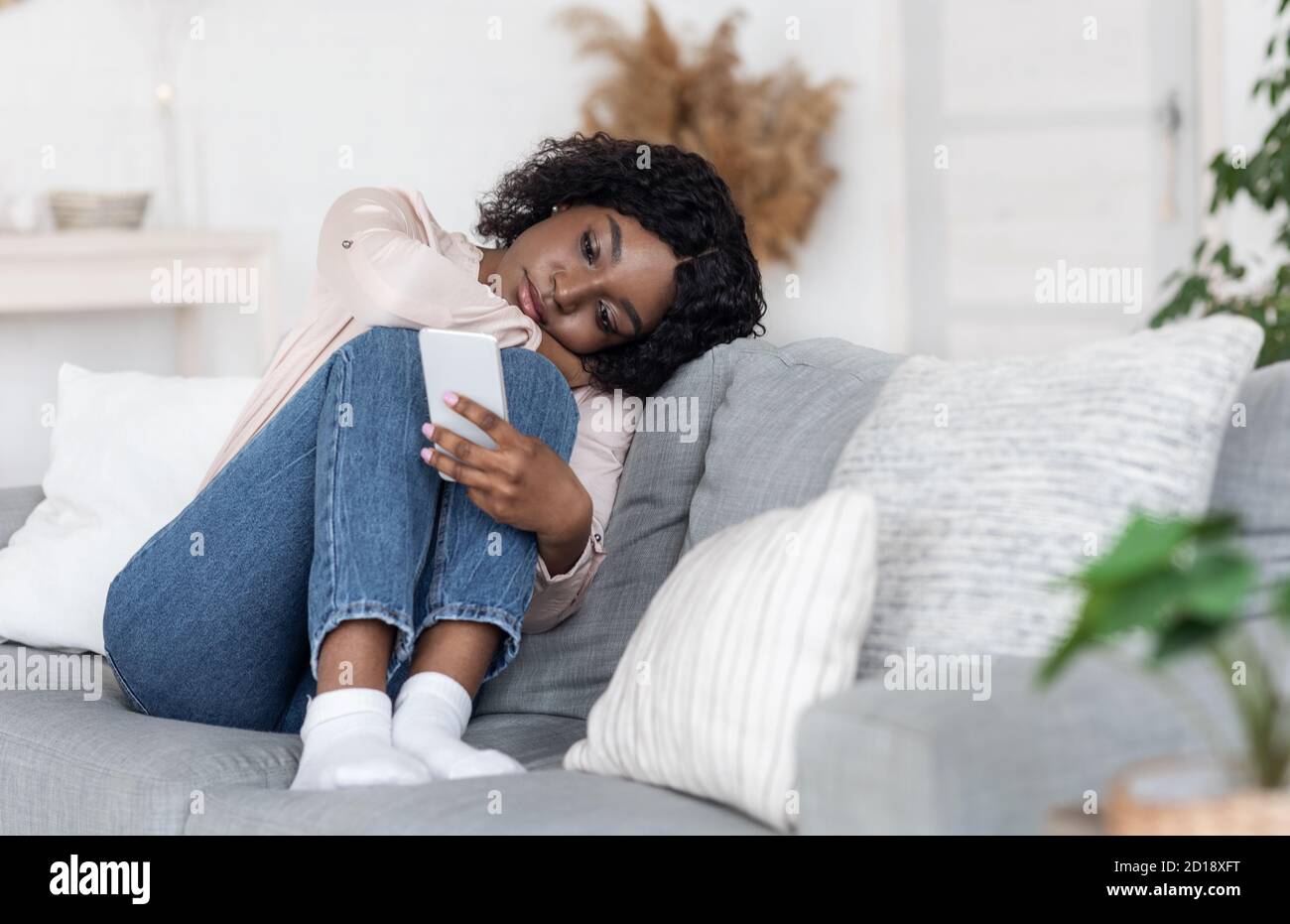 Einsamkeit. Depressive Schwarze Dame Sitzt Auf Der Couch Mit Smartphone, Loking Am Bildschirm Stockfoto