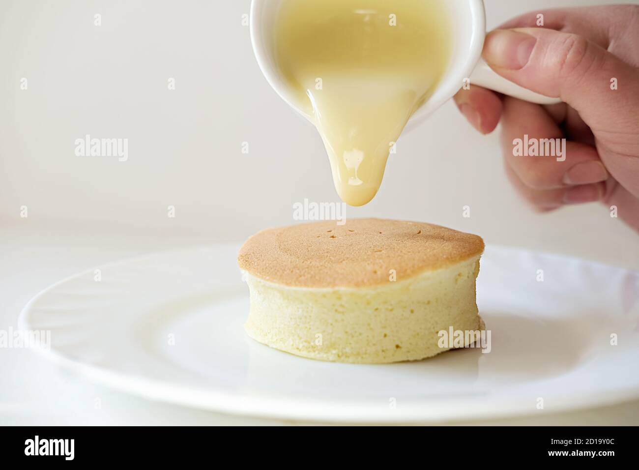 Trendiges asiatisches Frühstück, flauschiger japanischer Soufflepfannkuchen, Hand hält Würstchen und gießt Sahne auf heißen Pfannkuchen auf einem Teller Stockfoto