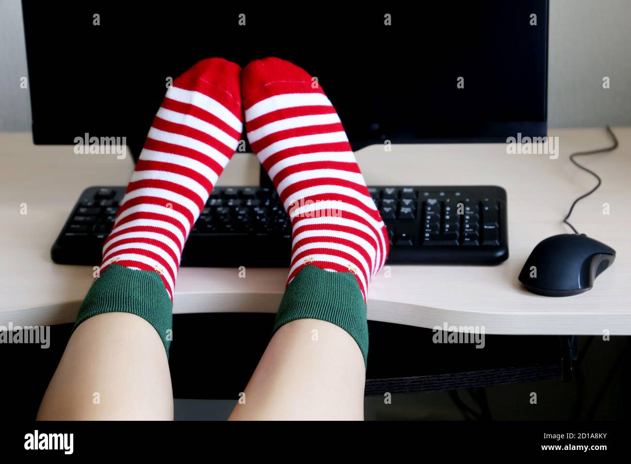 Weibliche Beine in Weihnachtssocken auf dem Schreibtisch mit PC und Tastatur. Konzept der Neujahrsfeier, Elf Kostüm Stockfoto