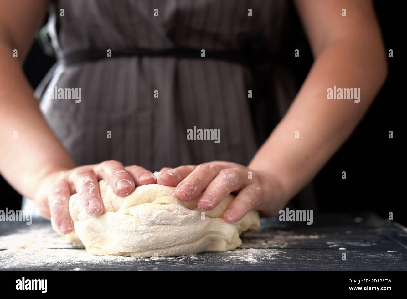 Frau Koch Kneten Teig für Brot oder Pizza. Bäckerei oder hausgemachtes Brot in Prozess, Frau in grau Schürze vor schwarzem Hintergrund. Konzept von Backen und Patisserie. Stockfoto