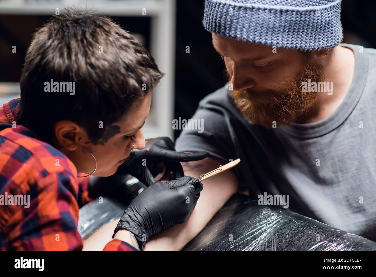 Ein Tattoo-Künstler mit kurzen Haaren und Piercings bereitet sich darauf vor, ein Tattoo auf den Arm eines Mannes in einem Tattoo-Salon zu bekommen. Stockfoto