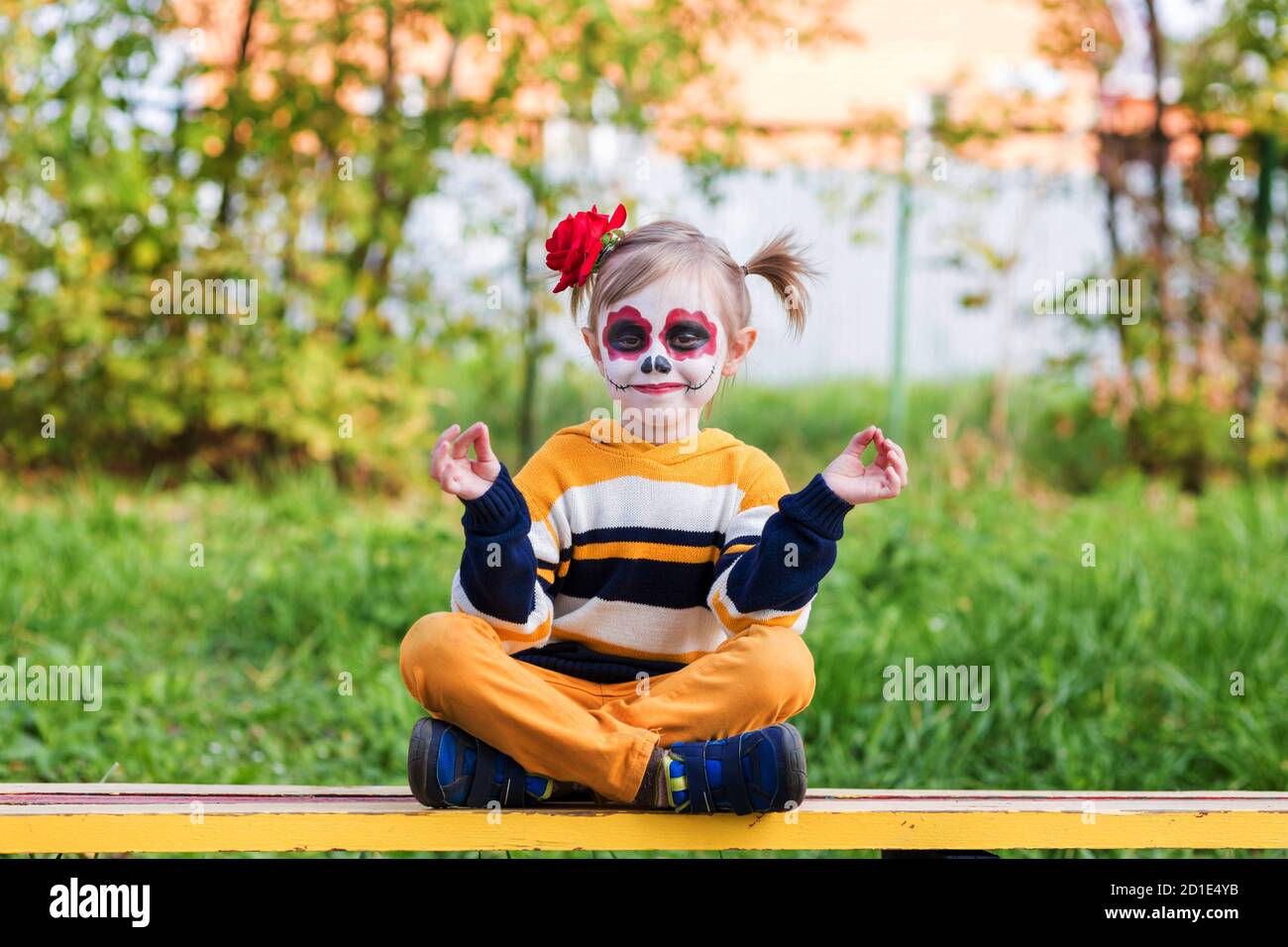 Ein kleines Mädchen mit gemaltem Gesicht, sitzend auf einer Bank am Tag der Toten. Stockfoto