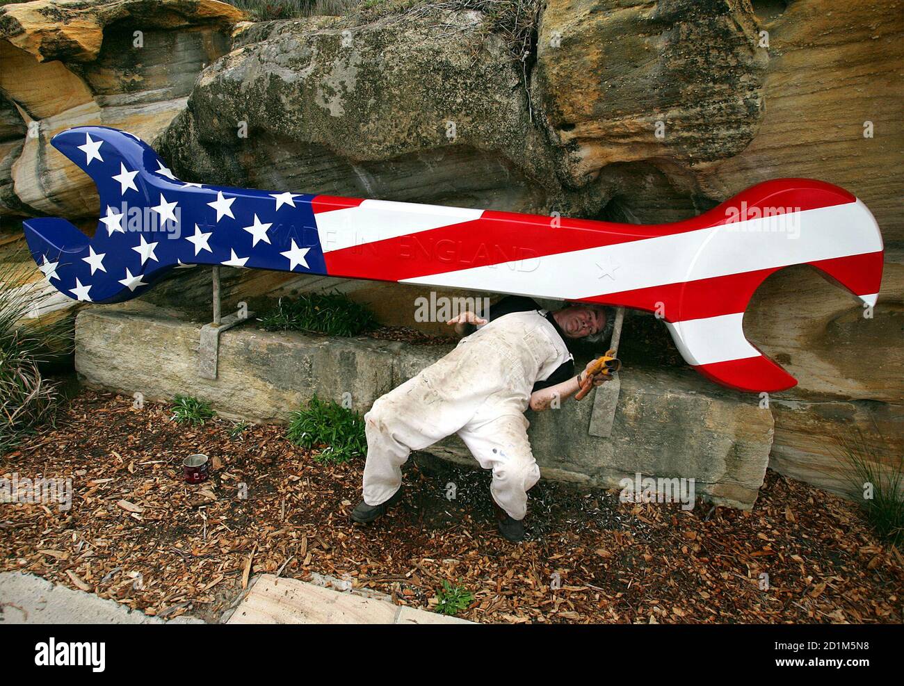 Künstler Peter Adams erleichtert heraus von unterhalb seiner Kunstwerke "A Star Bangled Sand" genannt, nach der Installation auf einer Klippe in der Nähe von Bondi Beach in Sydney Novembr 1, 2005. Seine Kunstwerke, aus Holz, Fiberglas, Harz, Stahl und Holz, stellt die USA Einmischung im Weltgeschehen, Unruhe weiterhin, wie "Sand im Getriebe", sagt Adams. Die Arbeit ist eine der über über 100 Exponate aus australischen und internationalen Künstlern, die Teilnahme an der jährlichen "Skulptur am Meer" Ereignis, das im neunten Jahr. Stockfoto