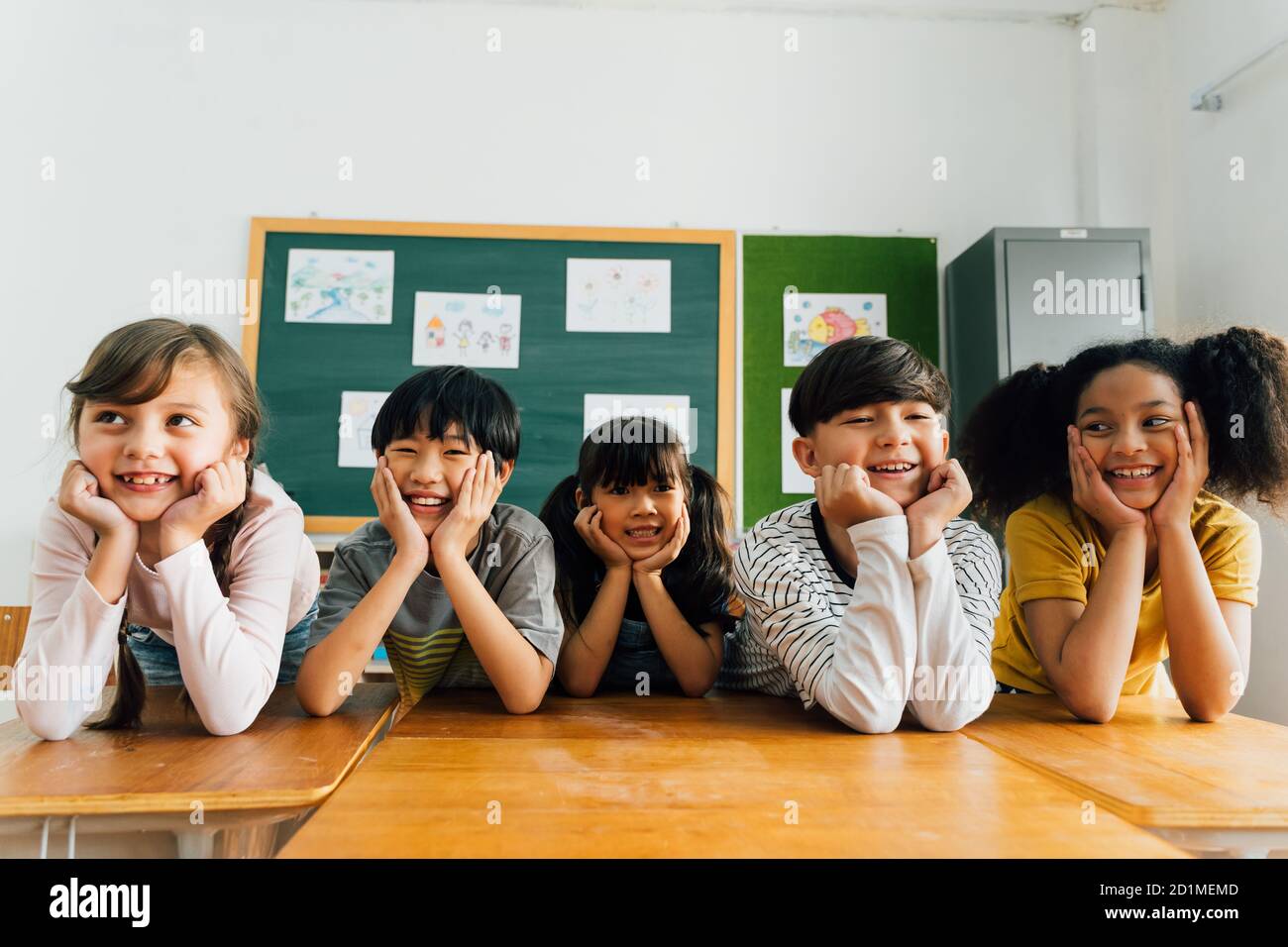 Fröhliche multiethnische Schulfreunde vor Kamera mit Händen am Kinn, Glück, Zweisamkeit, Freundschaft. Porträt von Schulkindern lächeln im Klassenzimmer Stockfoto