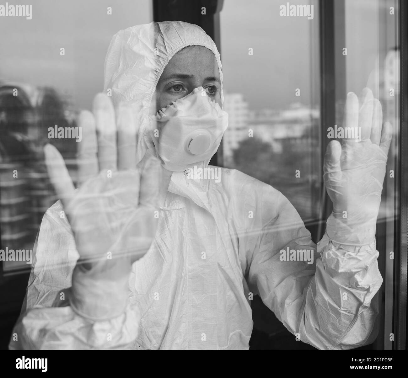 Porträt einer unglücklichen Ärztin, die PSA-Anzug, Atemschutzmaske, Handschuhe zur Vorbeugung trägt und durch das Fenster im Krankenhaus schaut. COVID-19 Konzept Stockfoto