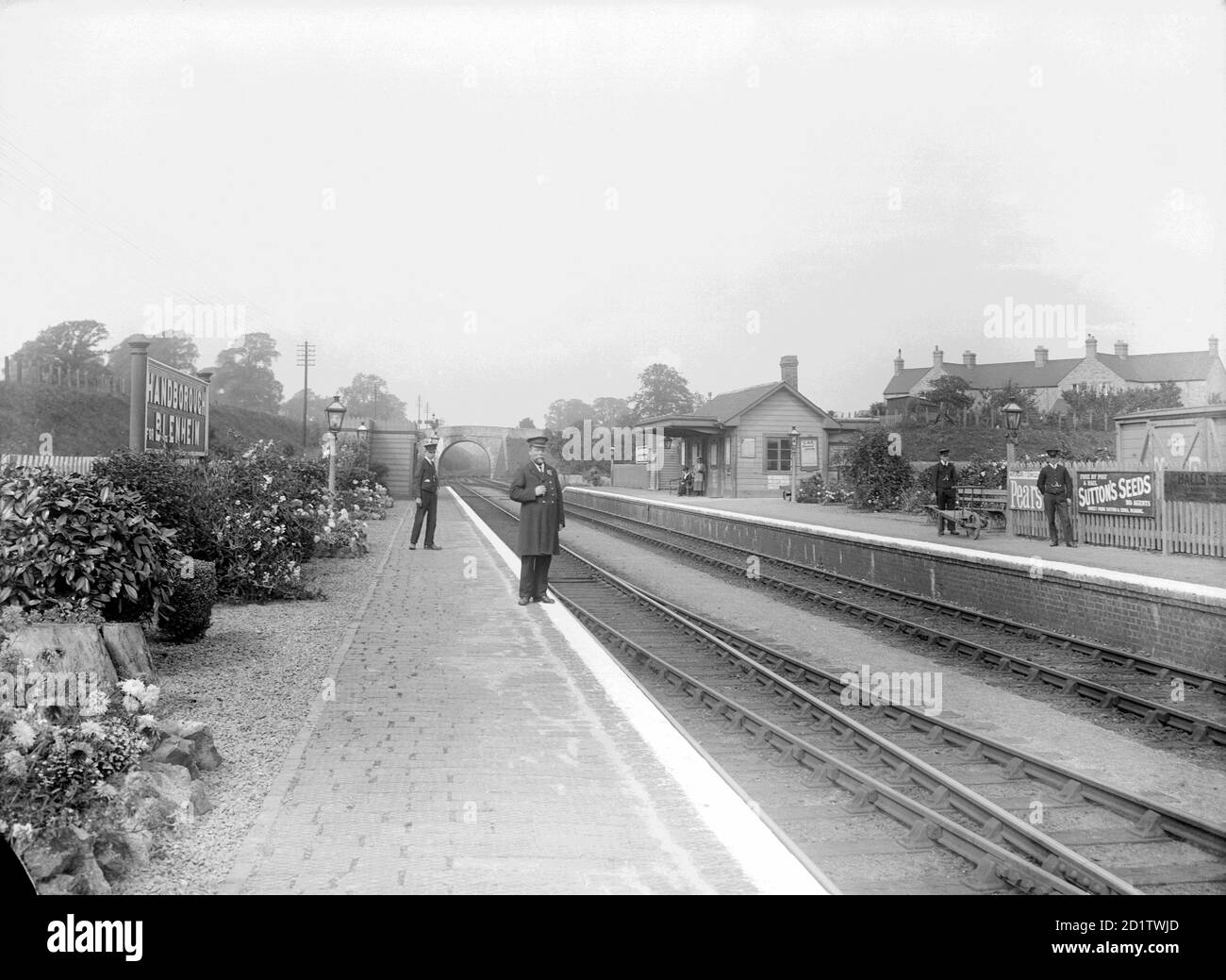 BAHNHOF LONG HANBOROUGH, West Oxfordshire. Der Bahnhofsmeister und eine Wache stehen auf der Plattform des kleinen Bahnhofs, mit einer Eisenbahnbrücke im Hintergrund sichtbar. Auf der gegenüberliegenden Plattform steht ein Portier mit Trolley. Fotografiert von Henry Taunt im Jahr 1920. Stockfoto