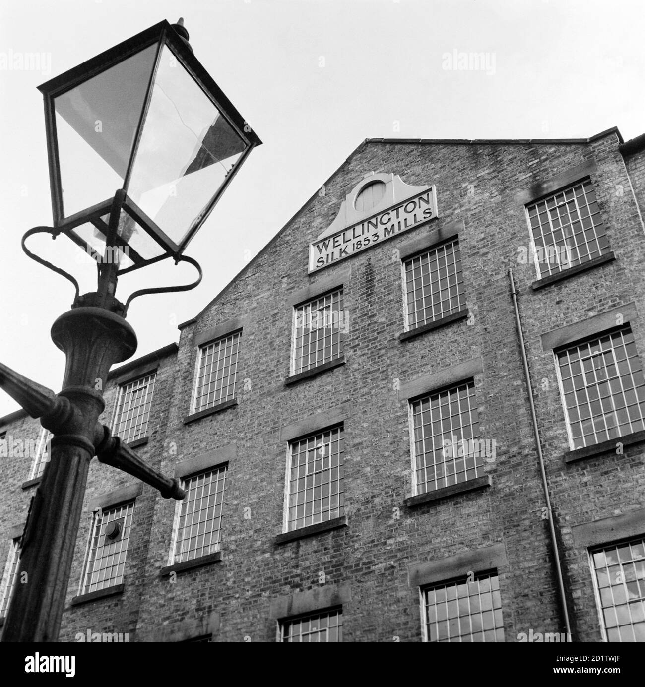 WELLINGTON MILLS, Strangman Street, Leek, Staffordshire. Außenansicht mit dem pedimented Abschnitt mit einer Lampe Standard im Vordergrund. Im 17. Und 18. Jahrhundert war Leek berühmt für die Herstellung von Seide von außergewöhnlicher Qualität. Als diese Mühle 1853 gebaut wurde, war der Import von französischer und italienischer Seide nach Großbritannien seit 25 Jahren illegal. Die Aufhebung dieses Verbots hatte katastrophale Auswirkungen auf die Seidenarbeiter in solchen Gebieten. Fotografiert von Eric de Mare im Jahr 1956. Stockfoto