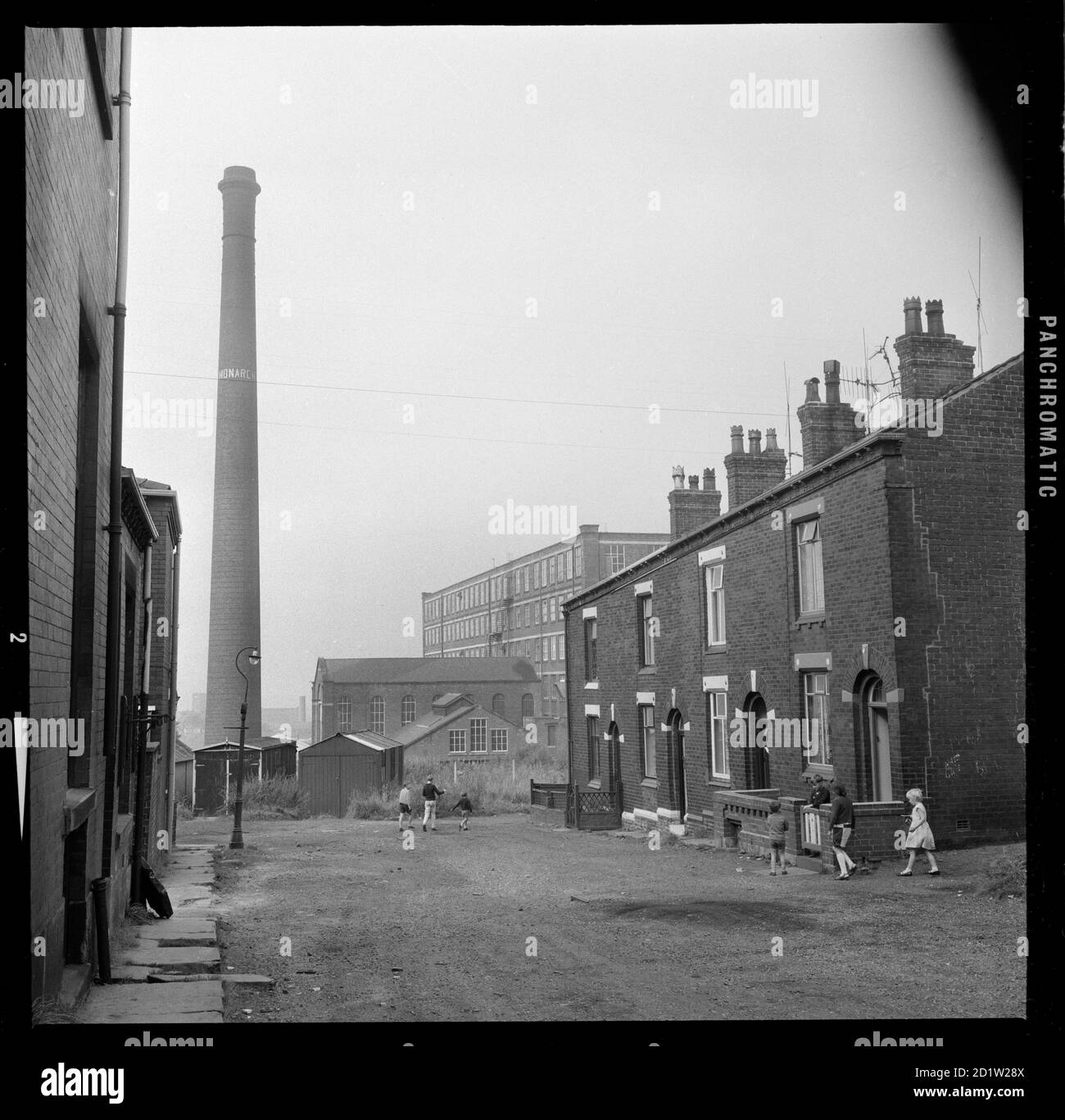 Das Äußere der Beswick Street 2-8 mit Monarch Mill im Hintergrund und einer Gruppe von Kindern außerhalb der Nummer 2, Oldham, Greater Manchester, UK. Stockfoto